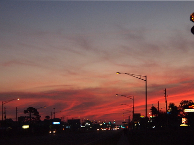 [Looking down a 6 lane roadway with the rising sun just off to the right. The street lights are still lit as are the other signs along the roadway. The sun has yet to reach the horizon so the sky is pink and grey close to the ground where the clouds are. The upper part of the sky is a white-blue as the sky begins lightening.]